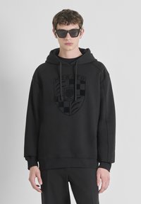 Sudadera negra con un logo frontal que presenta un patrón a cuadros y detalles de león. Tejido suave, capucha con cordón y un ajuste relajado. Combinada con pantalones negros.