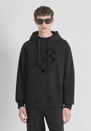 Haut noir à capuche avec un logo sur le devant présentant un motif à carreaux et des détails de lion. Tissu doux, capuche avec cordon et coupe décontractée. Associé à un pantalon noir.