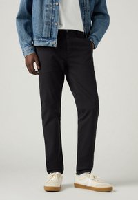 Pantalon noir en coton doux, avec une coupe droite et un style classique à cinq poches. Porté avec une veste en denim bleu et des baskets.