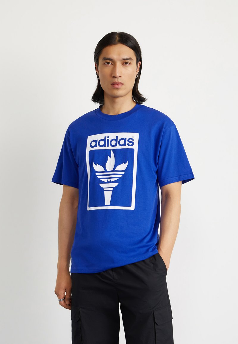Blue cotton t-shirt featuring a white Adidas logo with a torch design in the center. Short sleeves and crew neckline.