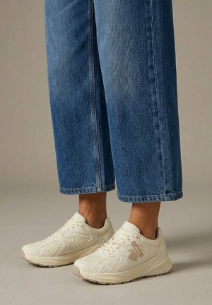 Gambe che indossano jeans corti in denim blu e sneakers chunky color crema con dettagli testurizzati, in piedi su una superficie liscia.