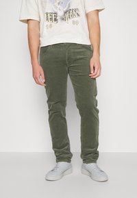 Pantaloni in velluto a coste verde oliva con vestibilità slim e superficie testurizzata, abbinati a sneakers bianche e una maglietta grafica color crema.