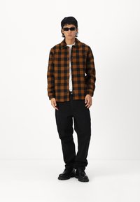 Brun og sort ternet flannel skjorte, sorte jeans og sorte støvler. Model iført solbriller, stående foran en ensfarvet hvid baggrund.