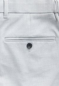 Pantalon gris clair ajusté avec une poche arrière fermée par un seul bouton noir et des coutures visibles sur une texture de tissu lisse.