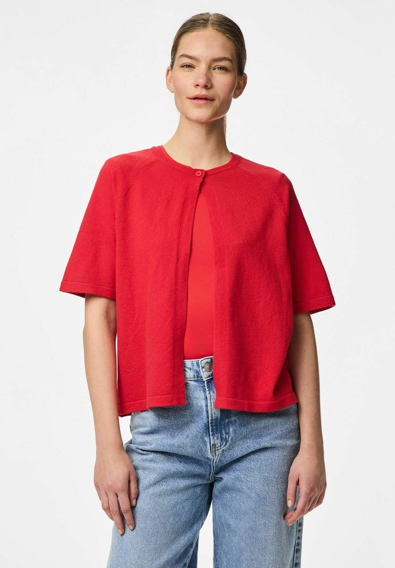 Crvena kratkotonna cardigan s kratkim rukavima, izrađena od mekanog materijala, s okruglim izrezom i jednim dugmetom na zatvaranju, u kombinaciji s plavim trapericama visokog struka.