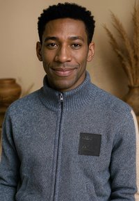 Grijze gebreide zip-up sweater met hoge kraag, voorzien van een gestructureerde stof en een zwart merkembleem op de borst.