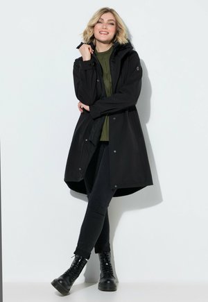 Jonge vrouw met blond haar, die een zwarte jas, groene top, zwarte broek en zwarte laarzen draagt, staande tegen een witte achtergrond.