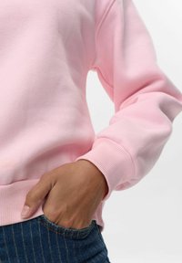 Rozā džemperis ar mīkstu tekstūru, ribotām piedurknēm un jostu. Roku atpūta zilajos svītrainajos džinsos, pabeidzot apģērbu.