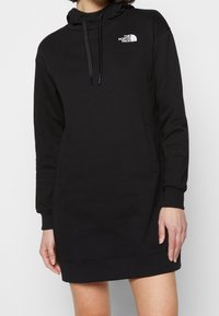 Svart hoodieklänning med en pulloverdesign, framficka och justerbara dragsko. Har en liten vit logotyp på bröstet.