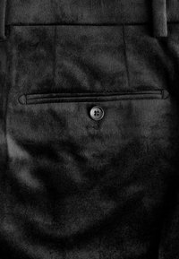 Tessuto scamosciato nero che mette in mostra una texture liscia, con un singolo bottone e un dettaglio nella tasca. Il design è minimalista e raffinato.