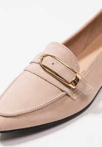 Beige suède mocassin met goudkleurige gesp, spitse neus en een gestructureerde bovenkant. Platte zwarte zool, subtiel gestikte accenten verbeteren het ontwerp.