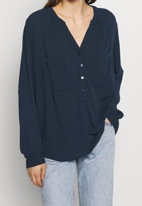 Donna che indossa una camicia blu scuro larga con bottoni e due tasche sul petto, abbinata a jeans azzurri, con una mano in tasca su uno sfondo neutro.