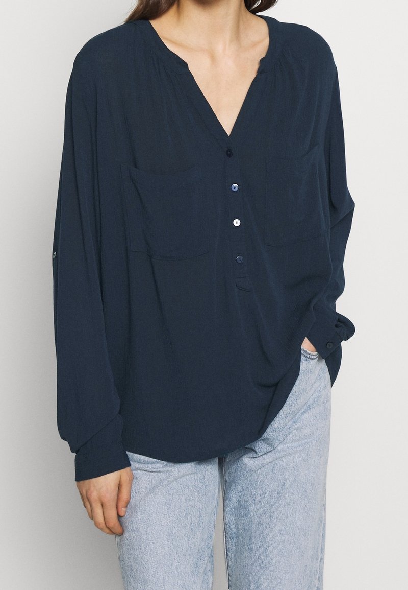 Donna che indossa una camicia blu scuro larga con bottoni e due tasche sul petto, abbinata a jeans azzurri, con una mano in tasca su uno sfondo neutro.