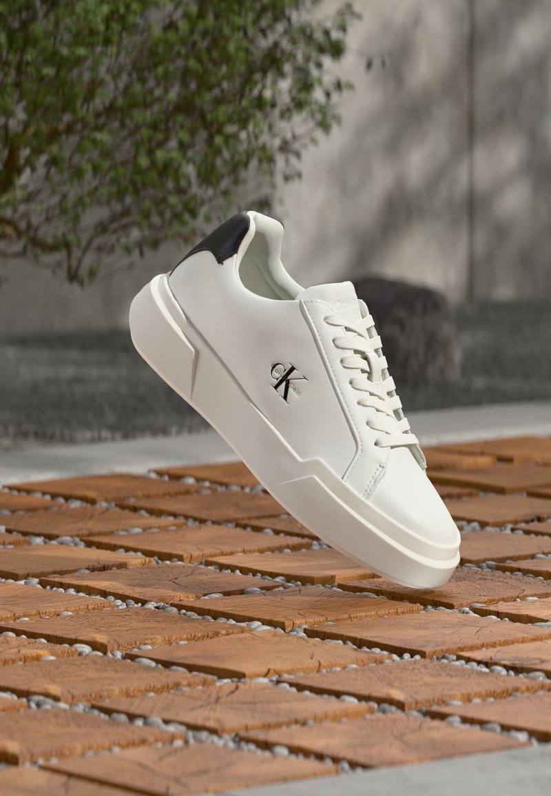 Λευκό χαμηλό αθλητικό παπούτσι με μαύρη λεπτομέρεια στη φτέρνα και λογότυπο CK, να αιωρείται πάνω από μονοπάτι από ξύλινα τούβλα με χαλίκια σε εξωτερικό χώρο.