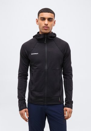 Άνδρας που φοράει μαύρο φούτερ με φερμουάρ και κουκούλα από την Mammut και μπλε σκούρο παντελόνι, στέκεται μπροστά σε απλό λευκό φόντο, κοιτάζοντας μπροστά.