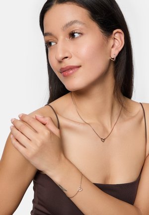 Roze-gouden sieradenset met hartvormige oorbellen, een delicaat ketting en een armband, gepresenteerd op een model met een zachte huid.