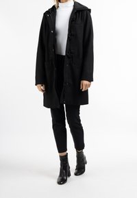 Czarny płaszcz o średniej długości z kapturem, z zapięciem na zamek, przednimi kieszeniami i zapięciem na napy. Nosi go na białym topie i czarnych spodniach.