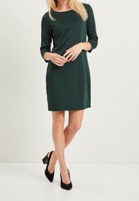 Robe vert foncé, longueur genoux avec un col rond et des manches trois-quarts, fabriquée en un tissu lisse et extensible. Assortie à des talons noirs.