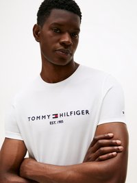 Λευκό βαμβακερό t-shirt με το λογότυπο "TOMMY HILFIGER EST. 1985" σε ναυτικό μπλε και κόκκινο. Στρογγυλή λαιμόκοψη, κοντά μανίκια, λεία υφή, εφαρμοστός σχεδιασμός.