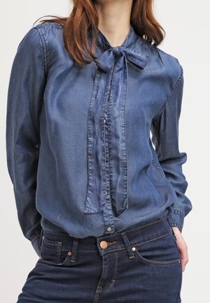 Frau trägt eine blaue Jeansbluse mit einer gebundenen Schleife am Kragen und dunkelblaue Jeans, steht mit einer Hand in der Tasche.