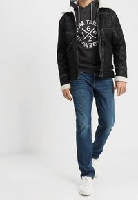 Zwarte suède jas met een schapenvacht kraag, grijze hoodie met een grafisch ontwerp, blauwe spijkerbroek en witte sneakers. Casual outfit.