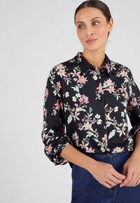 Blusa negra con un estampado floral en rosa, blanco y verde. Presenta un cuello con solapa y cierre de botón. Textura suave, mangas largas.