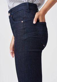 Dunkelblaue Jeans mit glatter Textur, hoher Taille und seitlichem Taschen-Detail. Mit einem Metallknopf und einem Logotag auf der Gesäßtasche.