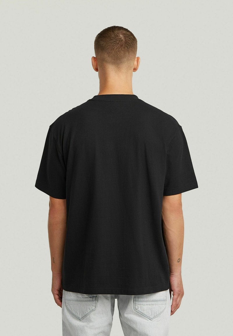 Uomo con capelli corti che indossa una maglietta nera semplice e jeans grigio chiaro, in piedi di schiena contro uno sfondo chiaro uniforme.