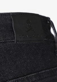 Un pantalon en denim noir présente une surface texturée, une patch en cuir avec un logo de skull et des coutures contrastantes, mettant en avant la durabilité et le style.