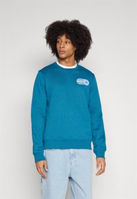 Teehl Sweatshirt mit gerippten Bündchen und Saum, ausgestattet mit einem blauen rechteckigen Logopatch auf der Brust. Über einem weißen Rundhals-Shirt getragen.