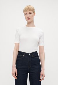 Camiseta ajustada blanca de manga corta, con cuello redondo, combinada con jeans de mezclilla oscuros de cintura alta que cuentan con cierre de botón y bolsillos frontales.