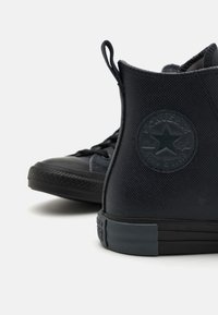 Crne visoke tenisice s gornjim dijelom od teksturiranog materijala, gumeni potplat i utisnuti Converse zvjezdasti logo sa strane. vezanje na vezice.