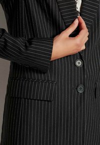 Schwarzer Blazer mit Nadelstreifen, der zwei schwarze Knöpfe, eine Vorderstasche und eine glatte Stoffoberfläche aufweist. Taillierte Passform mit strukturierten Schultern.