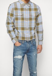Camisa de franela a cuadros en azul, verde y amarillo; con cuello abotonado y dos bolsillos en el pecho. Combinada con jeans azules desgastados.
