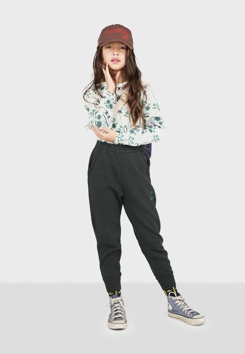 Sort joggingbukser med sidelommer, en mønstrede langærmet skjorte og en brun kasket. Grå og hvide sneakers med farverige sokker.