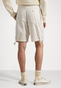 Muž v béžových šortkách na šňůrku, krémových ponožkách a béžových slip-on teniskách, stojící s rukama v kapsách, otočený zády.