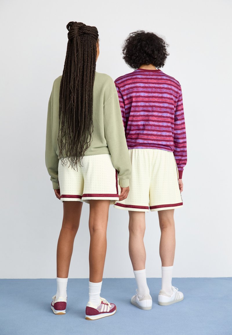 Deux personnes tournées le dos. L'une porte un pull surdimensionné vert clair, l'autre un haut rayé violet et rose. Les deux portent des shorts jaunes avec une bordure bordeaux.
