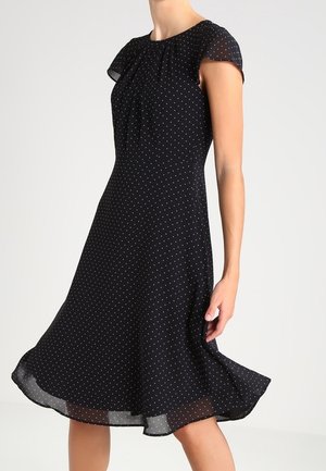 Femme portant une robe noire à petits pois blancs, manches courtes et ourlet évasé, se tenant devant un fond uni.