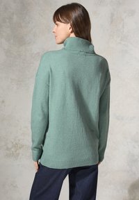 Mint green, oversized turtleneck sweater with ribbed accents at collar and hem, paired with dark blue loose-fitting pants. Soft texture.