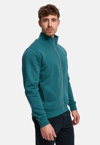 Teal genser med glidelås, høy krage, ribbede mansjetter og sidelommer. Stoffet har en glatt tekstur med en litt tilpasset passform.