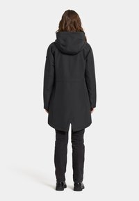 Svart vattentät jacka med huva, med en längre bakslut och en elegant design. Bärs över mörka byxor och svarta skor.