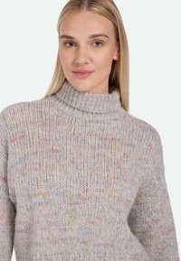 Light gray sweater with a high, chunky turtleneck. Knitted texture with multi-colored specks. Loose fit and dropped shoulder seams.