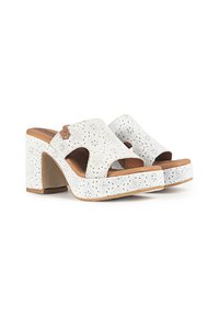 Mules blancos con tacón, con patrones decorativos en forma de flores recortadas, punta abierta y una suela de imitación de cuero en tono marrón. Tacón elevado para mayor estabilidad.