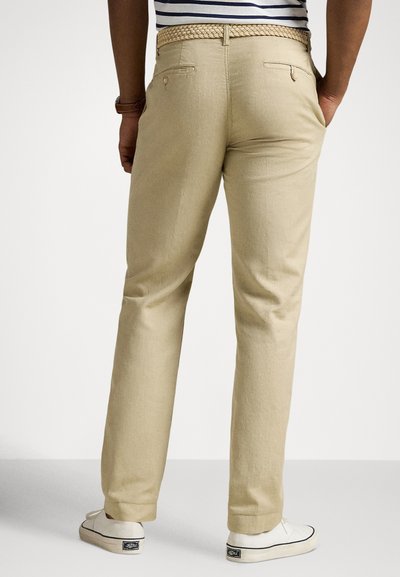 Bēšas krāsas činos bikses no viegla auduma ar taisnu kāju griezumu, austu jostu un aizmugurējo kabatu. Valkātas ar baltām sporta kurpēm.