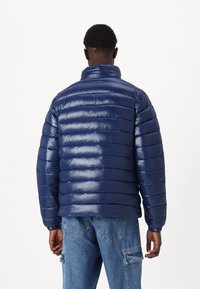 Marineblaues Puffer-Jackett mit glatter Textur, das mit genähten horizontalen Streifen, einem hohen Kragen und eng anliegenden Bündchen versehen ist. Getragen über Denim-Hosen.