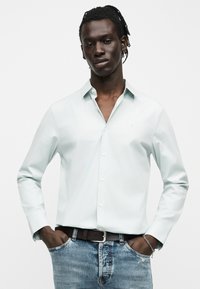 Νέος άνδρας που φορά ανοιχτό μπλε πουκάμισο με κουμπιά, μπλε τζιν, μαύρη ζώνη και ασημένιο βραχιολί, στέκεται με τα χέρια στις τσέπες.