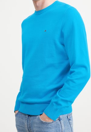 Uomo che indossa un pullover a maniche lunghe di un blu vivace con un piccolo logo sul petto, abbinato a jeans azzurri, con le mani nelle tasche.