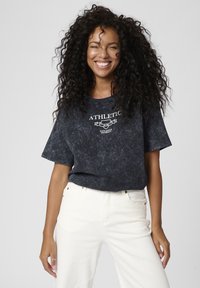 Dunkelgraues, kurzärmliges T-Shirt mit Rundhalsausschnitt und verblasster Optik, versehen mit weißem "ATHLETIC"-Text und Emblem-Druck. Kombiniert mit weißen Baumwolljeans.