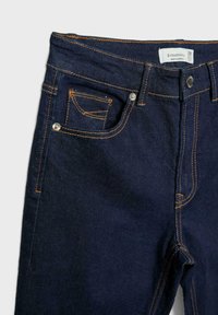 Temno modri denim kavbojki z kontrastnim oranžnim šivom. Značilnosti vključujejo standardni pas, sprednje žepe in kovinski gumb za zapiranje.