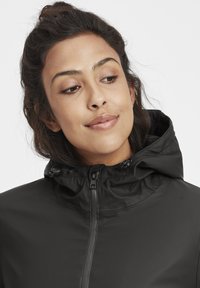 Chaqueta negra con capucha, de textura suave y mate, que cuenta con una cremallera frontal y cuerdas ajustables en la capucha para un ajuste personalizable.